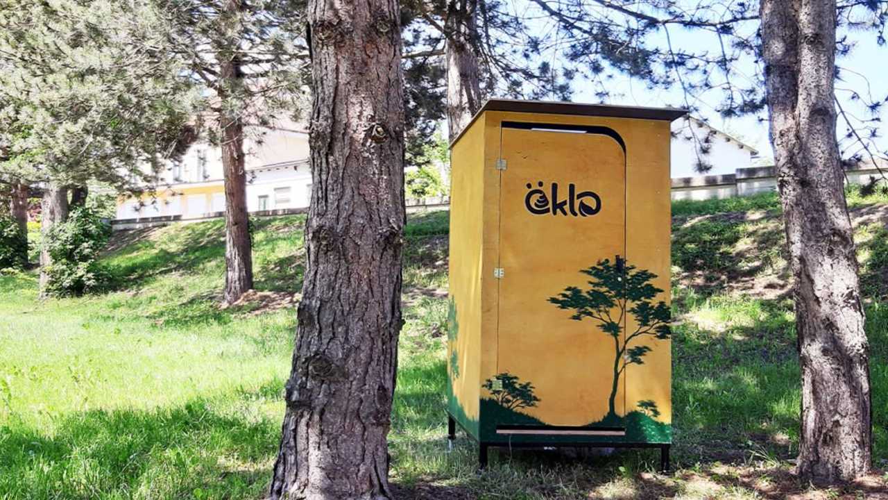 Ein öKlo mit grünem Baumaufdruck. Es steht zwischen zwei Bäumen, vor einem begrünten Abhang.