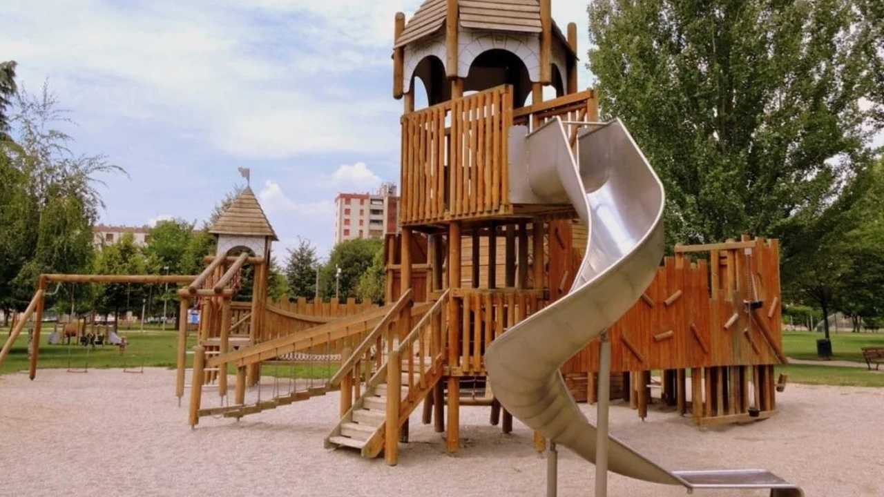 Eine Rutsche, die an einem Schloss-ähnlichen Gerüst auf einem Spielplatz angebracht ist. Das komplette Gerüst und die Türme sind aus Holz gebaut, die Rutsche ist aus Metall. Das ganze Gebilde ist auf einem Schotterfleck platziert.