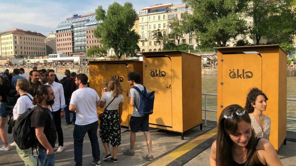 Drei öKlos, die entlang einer Flusspromenade stehen. Zwei Klassik und ein Barrierefrei-Modell sind zu sehen. Zahlreiche Menschen sind auf der Promenade davor und auf der anderen Seite des Flusses zu sehen.