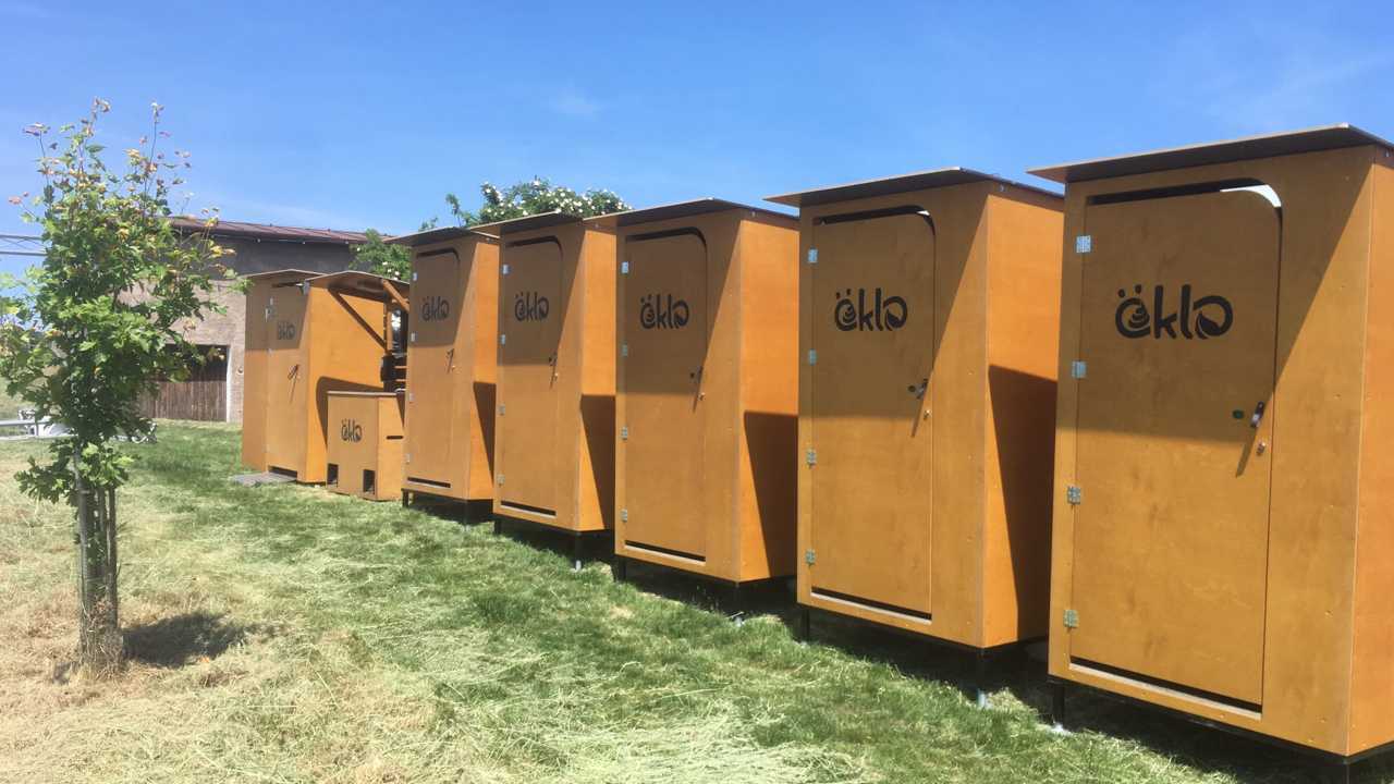 Einige öKlos in einer Reihe. An zweiter Stelle von links steht eine Waschstation zwischen den Toiletten. Alle öKlos sind auf einer grünen Wiese platziert, auf der auch ein kleiner Obstbaum wächst.