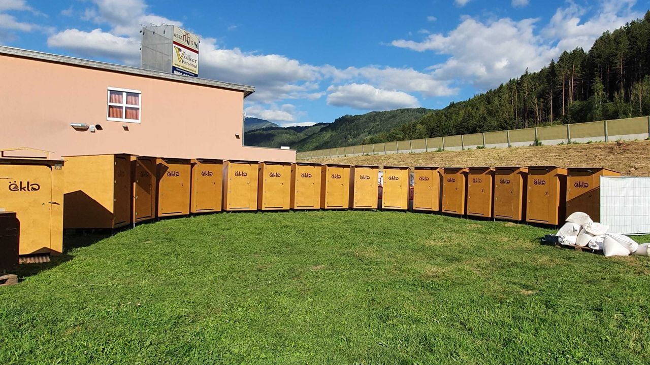 Eine Reihe öKlos, die auf einer grünen Wiese stehen. Dahinter ist ein rosa Haus zu sehen. Im Hintergrund erstreckt sich eine Bergkette unter blauem Himmel.