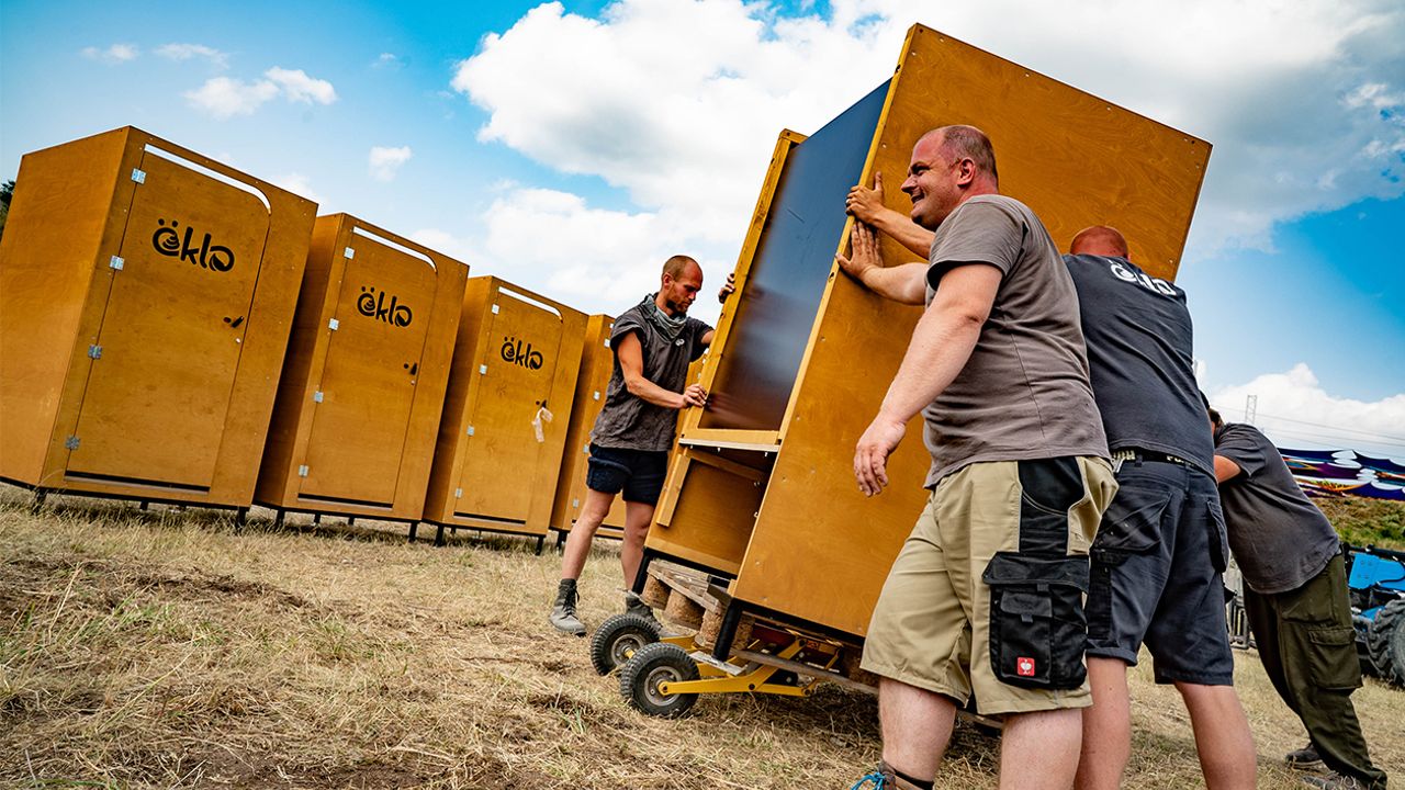 Vier Personen schieben eine öKlo-Kabine über einen Boden mit braunem, vertrocknetem Gras. Die Kabine steht auf einem kleinen Schubwagen.