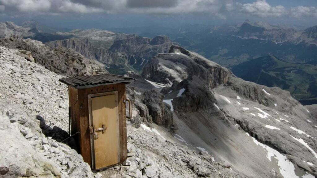 Eine Toilettenhütte aus Holz, die auf einem steilen Berghang inmitten der Steine steht. Sie ist mit einem Metallseil befestigt. Im Hintergrund sieht man aus der Vogelperspektive eine Bergkette, die sich weit erstreckt.