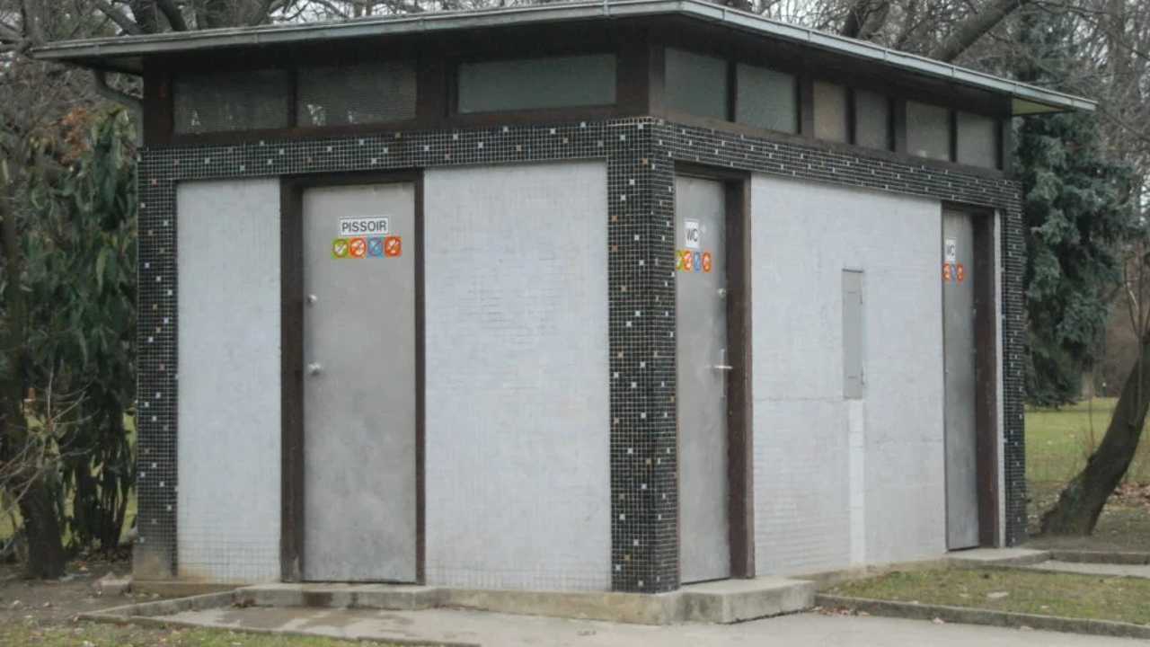 Eine öffentliche Toilettenanlage in einem Park, Die Wände sind grau. Auf den Türen an der rechten Seite ist die Aufschrift "WC" zu sehen, auf der linken Tür kann man die Aufschrift "Pissoir" sehen.