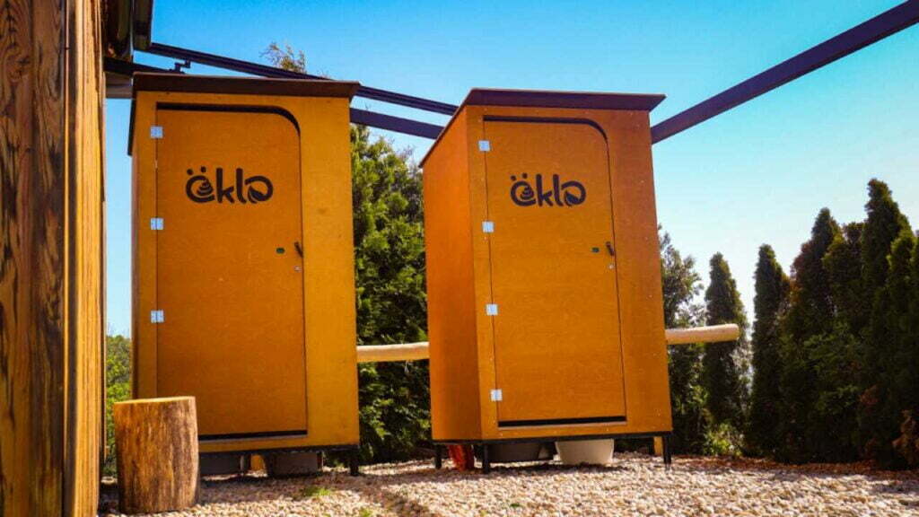 Zwei öKlo-Komposttoiletten, die nebeneinander vor einigem Bäumen stehen.
