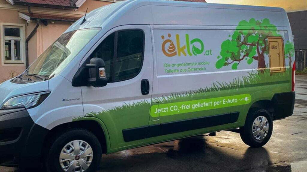 Ein E-Transporter, auf dessen Seite das öKlo-Logo zusammen mit dekorativen Elementen gedruckt ist. Der Slogan "Die angenehmste mobile Toilette aus Österreich" ist unter dem Logo zu sehen, ebenso wie die Aufschrift "Jetzt CO2-frei geliefert per E-Auto" mit einem Steckersymbol dahinter.