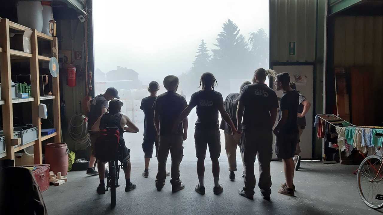 Neun öKlo-Mitarbeiter stehen vor den Toren der Halle. Diese sind geöffnet. Man sieht außerhalb nur vage Schemen von Bäumen, da starker Nebel herrscht.