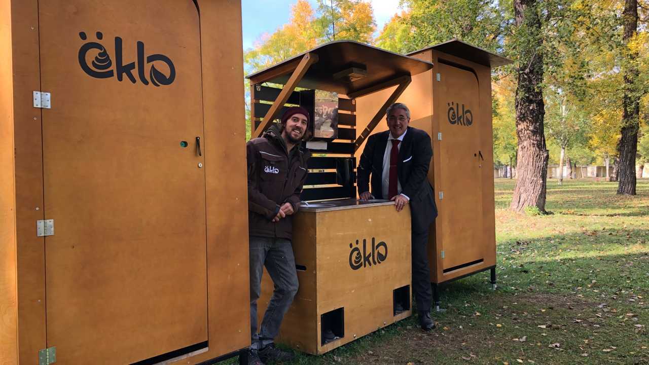 Zwei öKlo-Kabinen, in deren Mitte eine Waschstation steht. An die Waschstation sind Christian Schöner, einer der Gründer von öKlo, sowie ein Mieter unserer Toiletten gelehnt.
