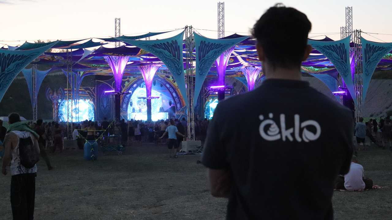 Ein Mann mit öKlo-Shirt steht abseits einer Festival-Halle und sieht den Besuchern beim Tanzen zu. Man sieht die Bühne nur teils, da das grelle Licht diese in einen Schein hüllt.