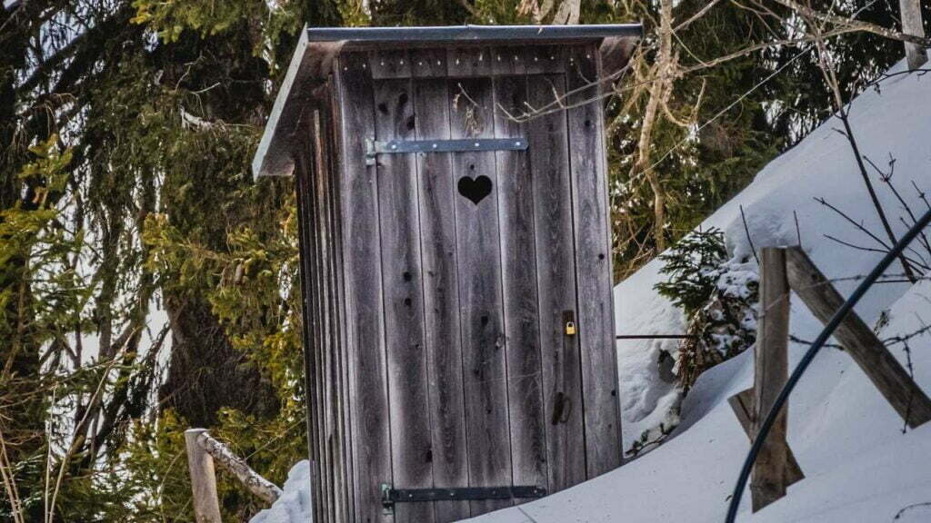 Eine klassiche Plumpsklo-Holzhütte mit einem Herzchen in der Tür. Um die Hütte herum liegt Schnee.