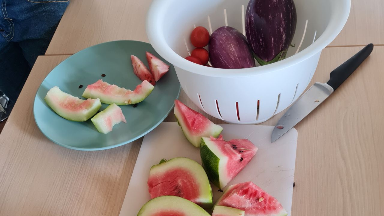 In Stücke geschnittene Wassermelone liegt auf einem Schneidbrett, das Messer direkt daneben. Ein Teller mit Schalen steht auf der anderen Seite. Darüber steht ein Sieb auf dem Tisch, in dem man Melanzani und Tomaten sehen kann.
