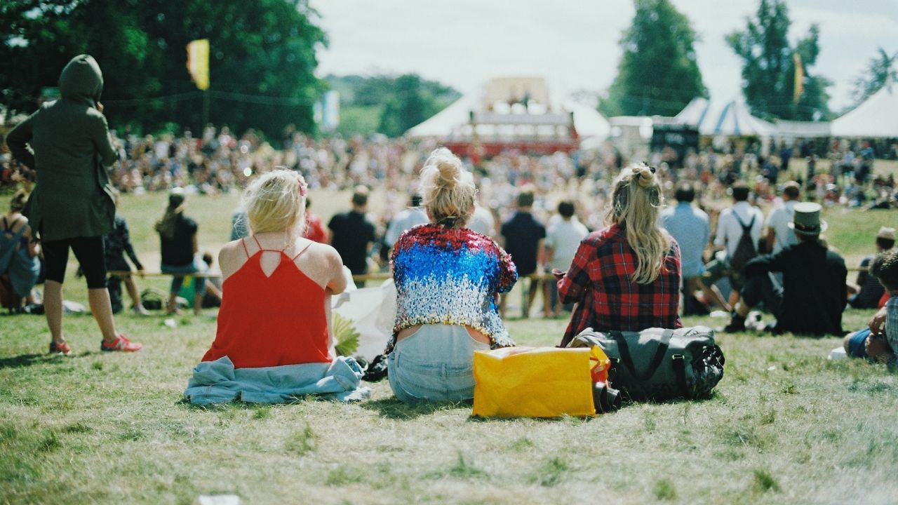 Drei Personen mit blonden Haaren sitzen mit dem Rücken zur Kamera. Die Menge vor ihnen sitzt, wie auch sie selbst, auf einer weiten Wiese. In der Ferne ist eine Bühne zu sehen. Vermutlich handelt es sich hierbei um ein Green Event.