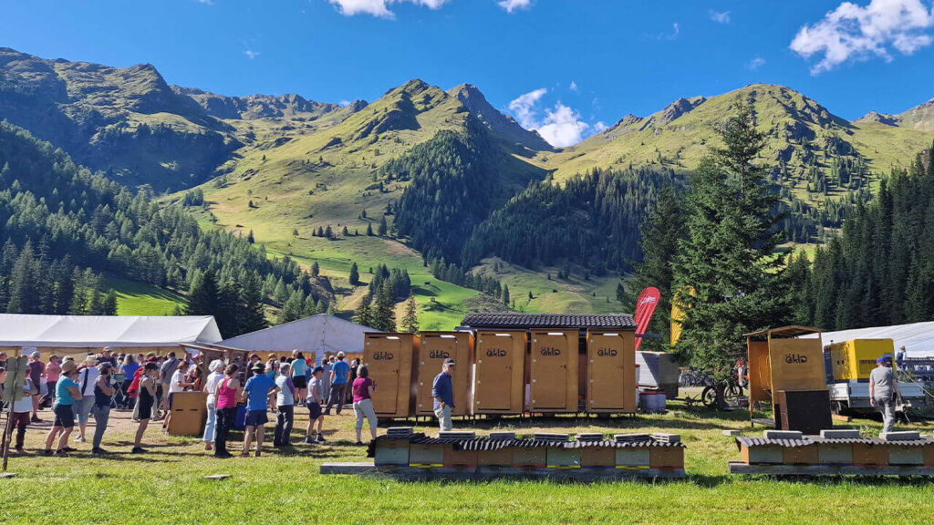 Eine Gruppe von Menschen steht in der Schlange vor mehreren öKlo-Komposttoiletten bei einem Outdoor-Event in den österreichischen Alpen mit grünen Bergen und klarem Himmel im Hintergrund.