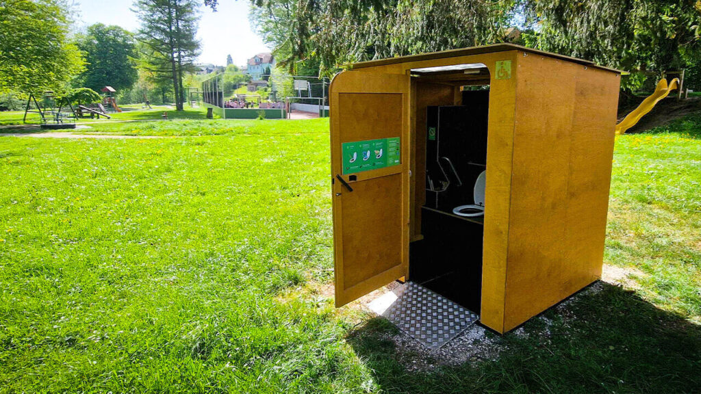 Eine Toilette von öKlo in einem Park mit viel Grün im Hintergrund, umgeben von Bäumen und einem Spielplatz.